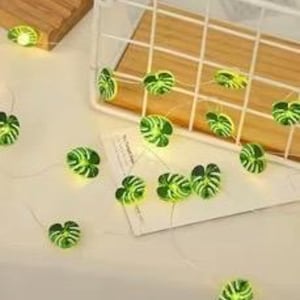 May include: String of LED lights with green, monstera-shaped covers. The lights are scattered on a white surface, with some placed inside a white wire basket and on a wooden block. The lights emit a soft, warm glow.
