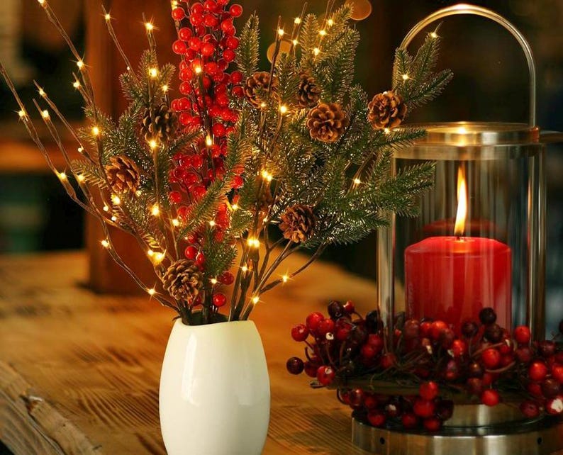 May include: A white vase with a twig arrangement featuring red berries and warm white LED lights. The vase is on a wooden table next to a glass lantern with a red candle and red berries.