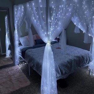May include: A white canopy bed with a white sheer curtain draped over it. The curtain is decorated with white fairy lights. The bed has a gray duvet and white pillows.