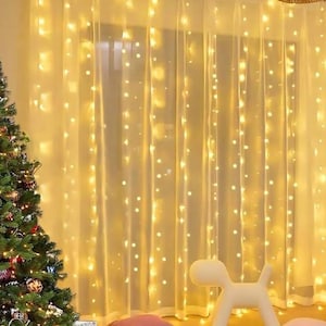 May include: A festive scene with a Christmas tree adorned with ornaments and wrapped gifts. Sheer curtains are illuminated by warm, twinkling fairy lights, creating a cozy ambiance. A decorative dog-shaped object and pillows are on the floor.