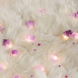 May include: A string of fairy lights with purple crystal-shaped lights on a white, fluffy surface. The lights are strung on a thin silver wire, and small pink flower-shaped accents are scattered around the crystals.