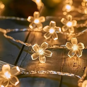 May include: Close-up of a string of fairy lights with clear, flower-shaped bulbs. The lights are on a thin, silver wire, and the bulbs are lit with a warm, yellow glow. The background is a dark wooden surface.