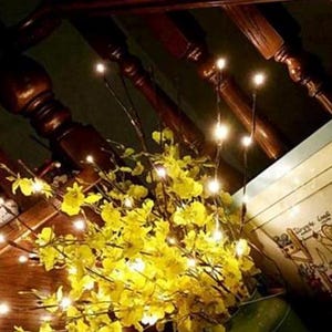May include: A set of warm white fairy lights strung on thin, flexible branches. The lights are clustered around a bouquet of yellow flowers, creating a warm and inviting ambiance.