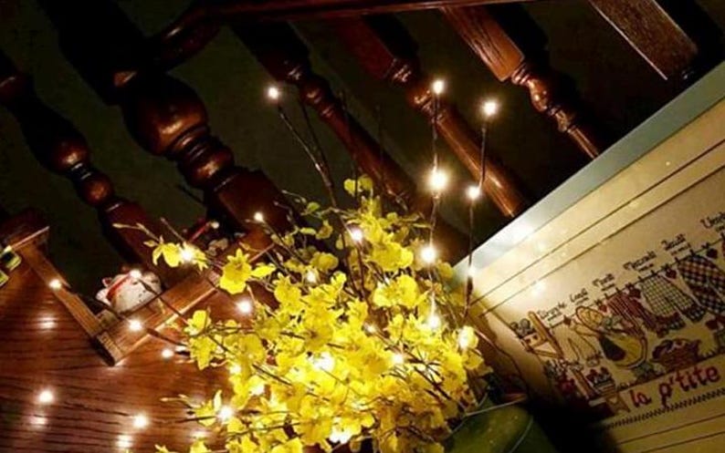 May include: A set of warm white fairy lights strung on thin, flexible branches. The lights are clustered around a bouquet of yellow flowers, creating a warm and inviting ambiance.