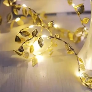 May include: A string of gold-colored leaf-shaped lights. The leaves are arranged along a thin, flexible wire, with small, bright lights interspersed among them. The lights are on, creating a warm, inviting glow. The overall effect is decorative and elegant.