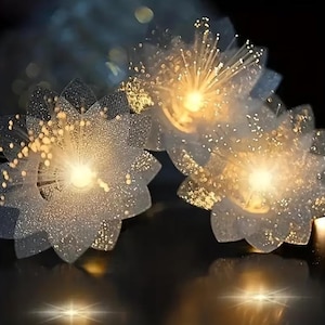 May include: Close-up of three illuminated, flower-shaped decorative lights. The lights have a white, glittery appearance with a central bright yellow glow and radiating fiber optic strands. The background is dark, creating a contrast with the lights.