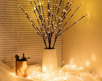 May include: A white ceramic vase with a decorative branch with warm white LED lights. The vase is sitting on a white surface with string lights.