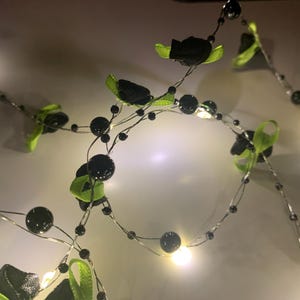 May include: A string of decorative lights with black beads and small black fabric flowers with green leaves. The lights are on a thin silver wire, creating a festive and elegant look. The lights are glowing.