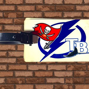 Può includere: Un'etichetta per bagagli bianca con un logo blu e bianco dei Tampa Bay Buccaneers e un logo rosso e bianco dei Tampa Bay Buccaneers. L'etichetta ha una cinghia per bagagli nera.