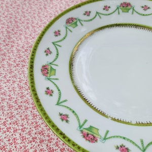 May include: A white porcelain plate with a green and gold rim. The plate is decorated with a floral pattern of pink roses and green leaves.