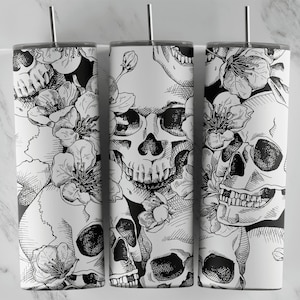 May include: White tumbler featuring a black and white floral skull design. The design includes detailed illustrations of skulls and blooming flowers. The tumbler has a silver lid and a matching straw. The overall aesthetic is gothic and artistic.