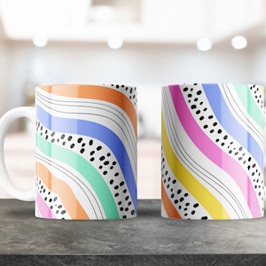 Puede incluir: Dos tazas de cerámica blanca con rayas onduladas de colores y lunares negros. Las tazas se enfrentan en una superficie gris.