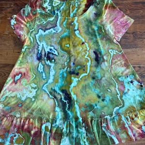 May include: A short-sleeve tunic top featuring a round neckline and a ruffled hem. The fabric displays a multi-coloured tie-dye pattern with swirling organic shapes in shades of olive green, turquoise, mustard yellow, deep purple, and soft pink.