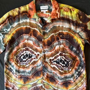 Könnte beinhalten: Ein kurzärmeliges Hemd mit Knopfleiste und einem Tie-Dye-Geodenmuster in Braun-, Orange-, Gelb- und Schwarztönen. Das Hemd hat einen Kragen, eine Brusttasche und ein Etikett mit der Aufschrift Saddlebred Classic Fit. Der Stoff zeigt konzentrische Ringe.