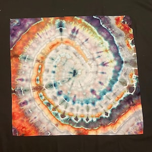 Może przedstawiać: Kwadratowy materiał tie-dye ze spiralnym wzorem. Wzór zawiera koncentryczne pierścienie kolorów, w tym pomarańczowy, fioletowy, turkusowy i biały. Tkanina jest ułożona płasko na czarnym tle.