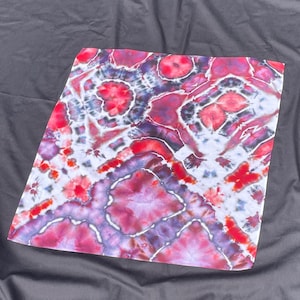 Może przedstawiać: Kwadratowy materiał tie-dye z żywym wzorem. Projekt zawiera mieszankę odcieni czerwieni, fioletu i bieli, tworząc efekt wizualny i artystyczny. Tkanina wydaje się miękka i elastyczna, odpowiednia do różnych zastosowań.