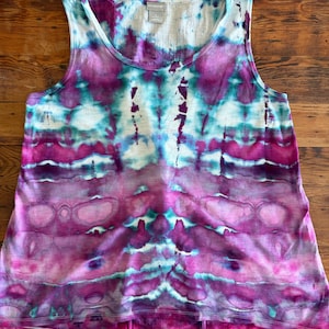 May include: A sleeveless tank top featuring a vibrant tie-dye pattern in shades of magenta, purple, and turquoise. The garment has a round neckline and a slightly flared hemline, with a white interior label visible at the inner back collar.