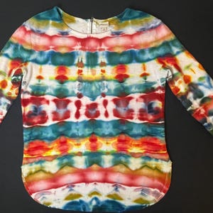 May include: A long-sleeved, scoop-neck shirt with a vibrant tie-dye pattern in red, orange, yellow, green, and blue. The shirt has a zipper at the neckline and a curved hemline. The sleeves are three-quarter length.