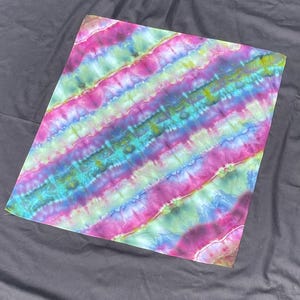 Może przedstawiać: Kwadratowy materiał tie-dye z żywym, ukośnym wzorem w paski. Projekt zawiera pasma w odcieniach różu, fioletu, niebieskiego, zielonego i żółtego. Tkanina wydaje się miękka i elastyczna, odpowiednia do różnych projektów rzemieślniczych lub odzieży.