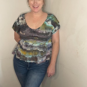 Op de afbeelding: Een tie-dye top met een gerimpelde zoom en korte mouwen. De top is in tinten blauw, groen, paars en bruin.