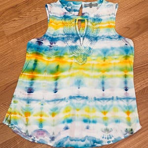 May include: Sleeveless tie-dye top in shades of blue, yellow, and white. The top features a keyhole neckline with a white crocheted lace detail. The fabric has a flowing, lightweight appearance, suitable for warm weather.