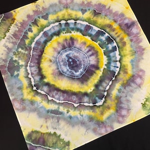 Może przedstawiać: Kwadratowy materiał tie-dye z koncentrycznymi okręgami w odcieniach żółtego, fioletowego, zielonego i niebieskiego. Wzór ma spiralny wzór z jasnożółtym tłem i ciemną czarną powierzchnią.
