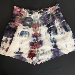 Op de afbeelding: High-waisted shorts met een tie-dye patroon in tinten wit, paars, blauw en kastanjebruin. De shorts hebben een geplooide voorkant en een geplooide tailleband. De stof lijkt licht van gewicht.