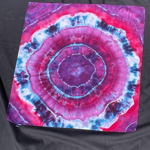 Może przedstawiać: Kwadratowy materiał tekstylny tie-dye z wzorem koncentrycznych okręgów. Wzór zawiera pierścienie w odcieniach fioletu, różu i niebieskiego. Tkanina jest prezentowana na czarnej powierzchni.