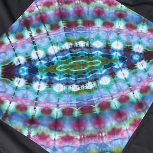 Może przedstawiać: Kwadratowy materiał tie-dye z żywym, wirującym wzorem. Projekt zawiera mieszankę kolorów, w tym niebieski, fioletowy, zielony i biały, tworząc efekt psychodeliczny. Tkanina jest umieszczona na czarnym tle.