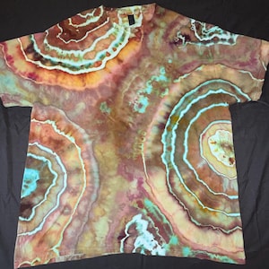 May include: A short-sleeve t-shirt featuring a multi-colored tie-dye pattern that mimics the concentric rings of a geode. The design incorporates shades of brown, tan, rust, and turquoise, with white lines separating the circular color bands.