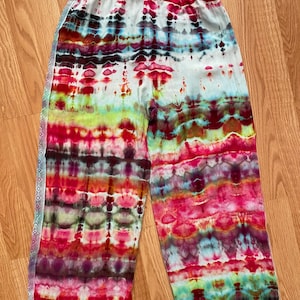 May include: Tie-dye wide-leg pants with a colorful, striped pattern in shades of red, pink, green, blue, and black. The pants have a white elastic waistband and a white lace trim along the sides.