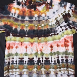 Könnte beinhalten: Ein kurzärmeliges Hemd mit Knopfleiste und einem horizontalen Batikmuster in Braun-, Orange-, Gelb-, Grün- und Weißtönen. Das Hemd hat einen Reverskragen, eine Knopfleiste mit fünf sichtbaren Knöpfen und vertikale Ziernähte in Form von Biesen.