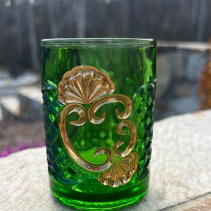 May include: A green glass tumbler with a gold-colored design. The design features a stylized floral motif with a scalloped edge.
