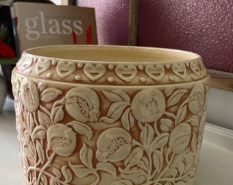 May include: A ceramic planter with a cream-colored glaze and a raised design of pomegranate branches. The planter has a slightly rounded shape and a wide rim.