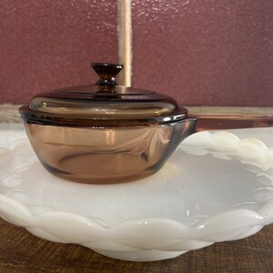 May include: A brown glass casserole dish with a lid and a handle. The dish has a floral design etched into the glass.