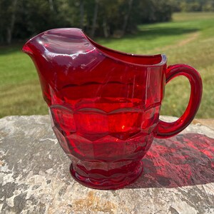 May include: A red glass pitcher with a textured, diamond-shaped pattern. The pitcher has a wide, rounded spout and a handle.