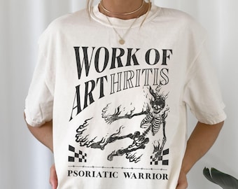 May include: Cream-colored t-shirt with a black graphic design. The design features the text "WORK OF ARTHRITIS" above a skeletal figure engulfed in flames, with the words "PSORIATIC WARRIOR" below. The shirt has short sleeves.
