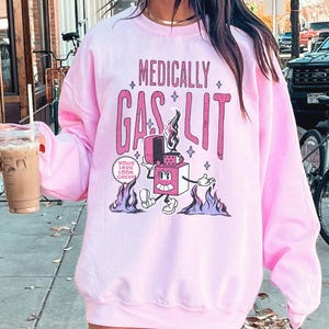 Pode incluir: Camisola cor-de-rosa com um gr&aacute;fico de um isqueiro de desenho animado com o texto "Medically Gaslit" e "Your Labs Look Great!" impresso nela.