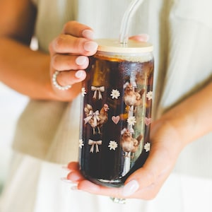 May include: A reusable glass cup with a brown lid and a clear straw. The cup is filled with a dark brown liquid and has a pattern of brown chickens, pink bows, and white flowers.