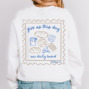 May include: White sweatshirt with a brown and blue graphic of a stamp with the text "give us this day our daily bread Matthew 6:11" and a drawing of bread and other food items.