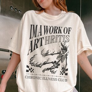 Pu&ograve; includere: Una maglietta color crema con una grafica nera di uno scheletro in fiamme. Il testo sulla maglietta recita "I'm a work of arthritis" e "Chronic Illness Club".