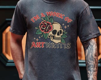 May include: A dark gray t-shirt with a vintage-style skull and rose graphic. The text "I'm a work of arthritis" is printed below the graphic.