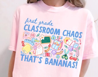 以下が含まれることがあります： 「first grade CLASSROOM CHAOS THAT'S BANANAS!」という青い文字がプリントされたライトピンクのTシャツ。定規、ハサミ、バックパック、黒板、A+の評価が書かれた紙など、カラフルな学用品のイラストが周囲に描かれています。