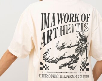 May include: A cream-colored t-shirt with a black graphic print of a skeleton on fire. The text "I'M A WORK OF ARTHRITIS CHRONIC ILLNESS CLUB" is printed on the shirt.