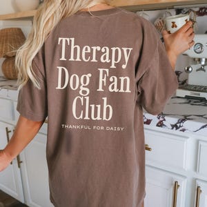 May include: A brown t-shirt featuring the words "Therapy Dog Fan Club" in white, with "THANKFUL FOR DAISY" underneath. The shirt appears to be a relaxed fit, possibly made from cotton or a similar material.