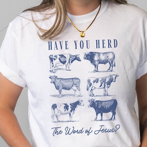 Peut inclure: T-shirt blanc avec un graphique bleu de vaches et le texte "Have You Herd The Word of Jesus?"
