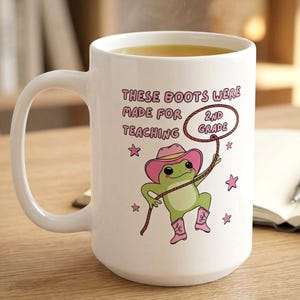 May include: White ceramic mug with a cartoon frog wearing a pink cowboy hat and boots, holding a lasso. Text on the mug reads "These boots were made for teaching 2nd grade" with pink stars.