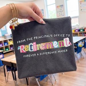 May include: A dark gray tote bag with the text "FROM THE PRINCIPAL'S OFFICE TO Retirement FOREVER A DIFFERENCE MAKER" in white and colorful letters. The bag is held in a classroom setting.