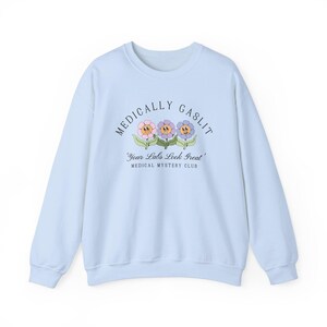 May include: Light blue sweatshirt with the text "Medically Gaslit" and a graphic of three cartoon flowers with faces. The text below the graphic reads "Your Labs Look Great" and "Medical Mystery Club".
