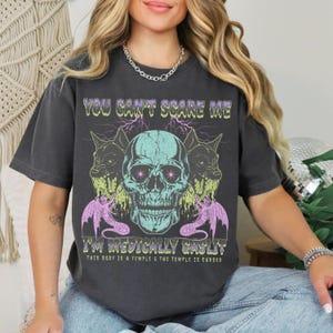 May include: Dark gray t-shirt with a graphic design featuring a teal skull, two wolf-like creatures, and text that reads "YOU CAN'T SCARE ME I'M MEDICALLY GASLIT. THIS BODY IS A TEMPLE & THE TEMPLE IS CURSED."